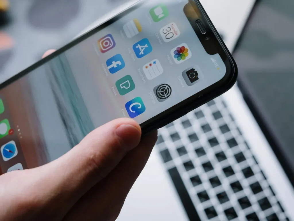 Hand holding a smartphone with apps in front of a laptop